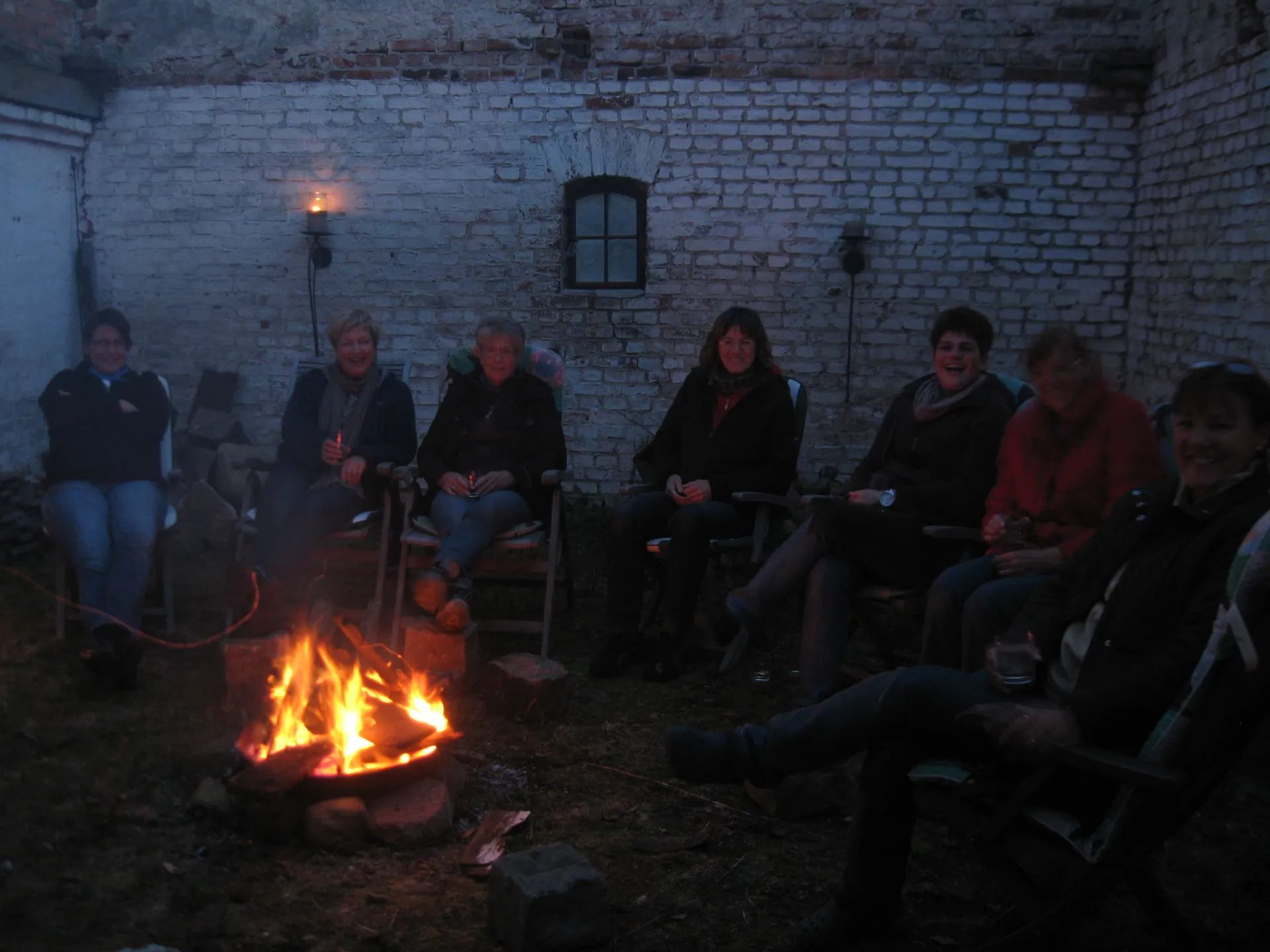 Gäste sitzen abends in der Runde am Lagerfeuer vor der Backsteinwand
